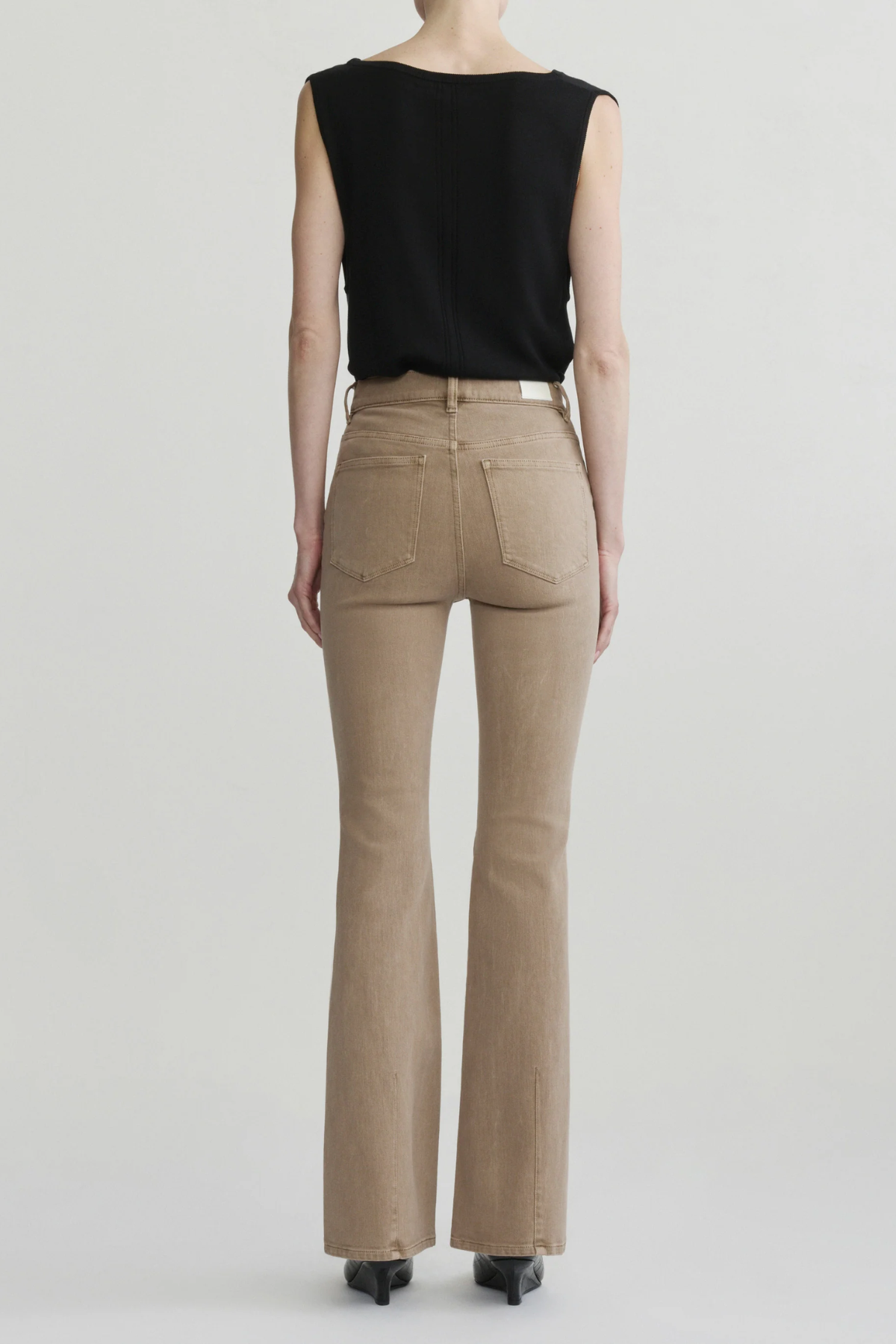 A model shown from the back wearing beige flared jeans, a black sleeveless top, and black heels against a light gray studio background.
