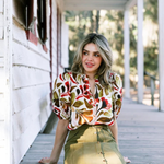 A woman sits on a wooden porch wearing a short-sleeve blouse with a bold multicolor abstract pattern in red, orange, green, and brown, paired with an olive green high-waisted skirt.