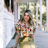 A woman sits on a wooden porch wearing a short-sleeve blouse with a bold multicolor abstract pattern in red, orange, green, and brown, paired with an olive green high-waisted skirt.