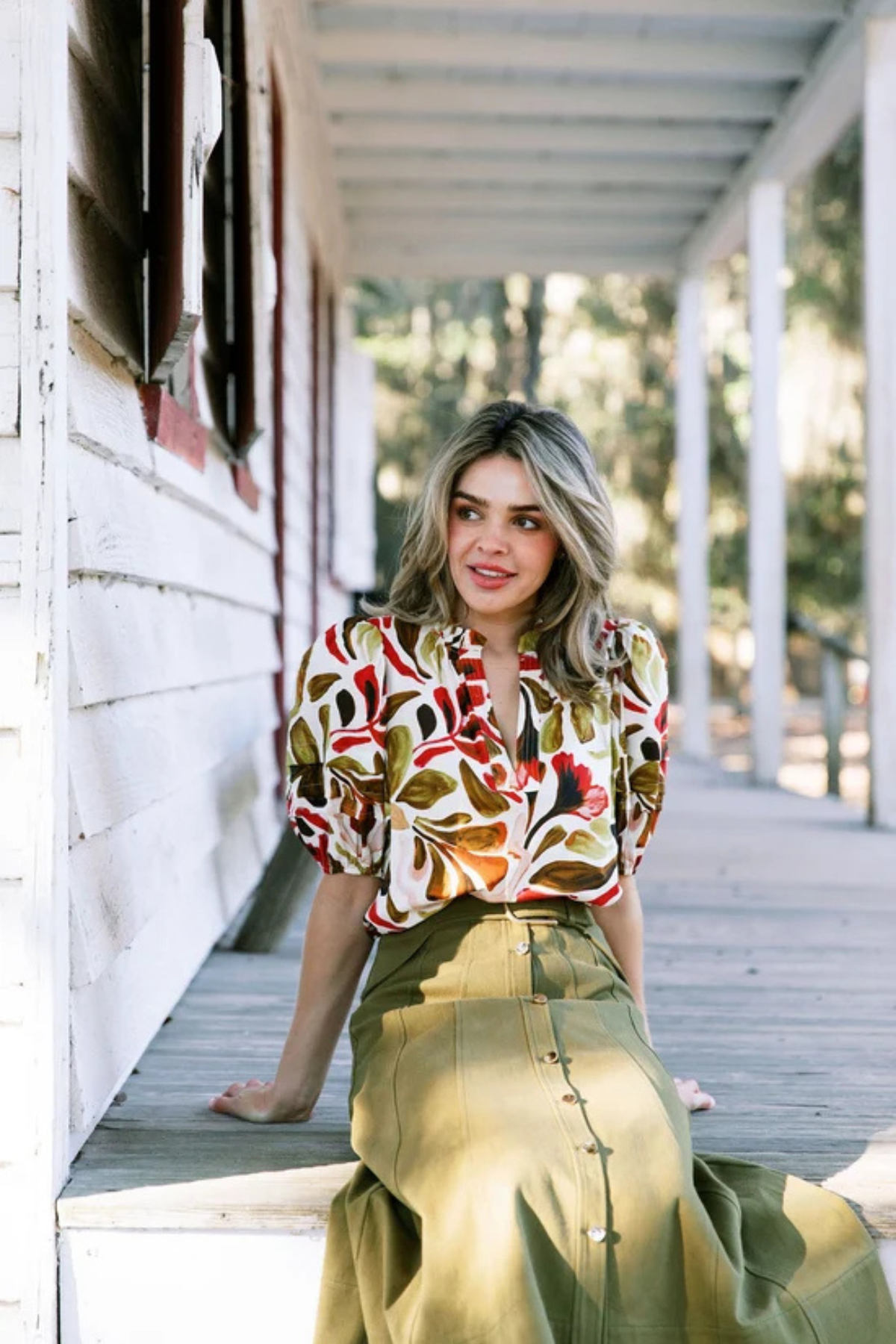 A woman sits on a wooden porch wearing a short-sleeve blouse with a bold multicolor abstract pattern in red, orange, green, and brown, paired with an olive green high-waisted skirt.