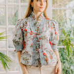 Woman wearing a floral blouse standing in front of a window with plants.