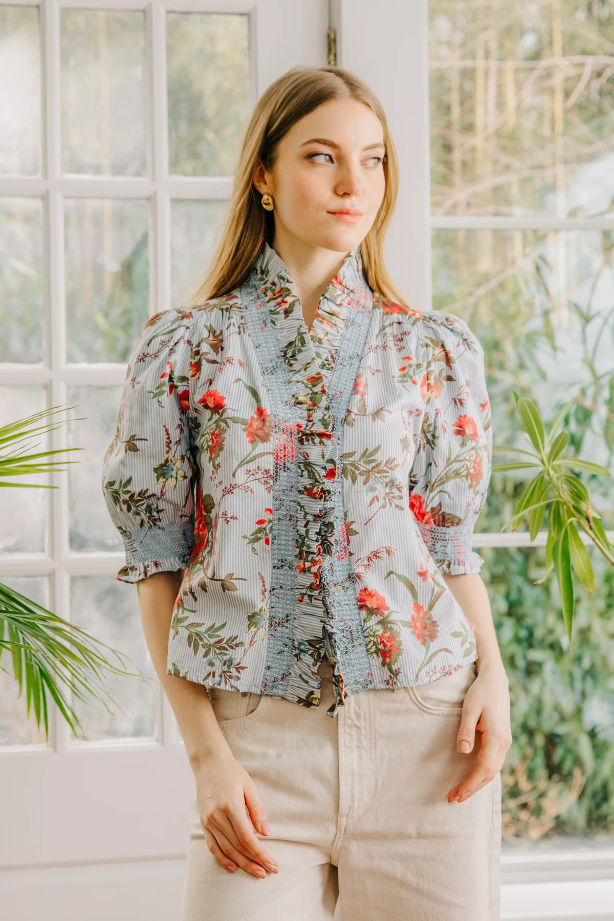 Woman wearing a floral blouse standing in front of a window with plants.