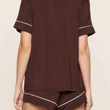 A woman wearing a dark brown short-sleeve pajama top and matching shorts with thin white piping trim, shown from the back against a light beige background.