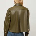 A woman viewed from the back wearing an olive green faux leather cropped jacket with gold-tone buttons on the cuffs, paired with blue jeans, against a light gray studio background.