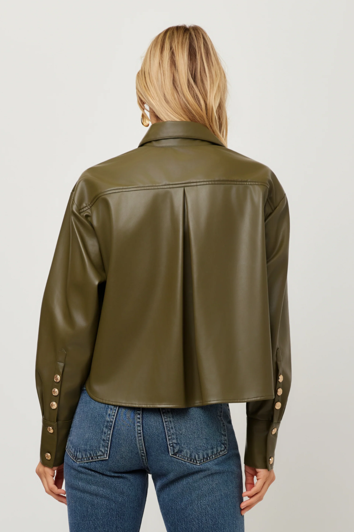 A woman viewed from the back wearing an olive green faux leather cropped jacket with gold-tone buttons on the cuffs, paired with blue jeans, against a light gray studio background.