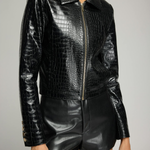 A model wearing a black crocodile-embossed faux-leather jacket with a front zipper and black faux-leather shorts, shown from the chin down against a neutral gray studio background.