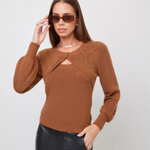 A woman wearing a brown ribbed knit long-sleeve sweater with a keyhole cutout at the chest, paired with black faux-leather pants, against a light gray studio background.