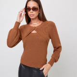 A woman wearing a brown ribbed knit long-sleeve sweater with a keyhole cutout at the chest, paired with black faux-leather pants, against a light gray studio background.