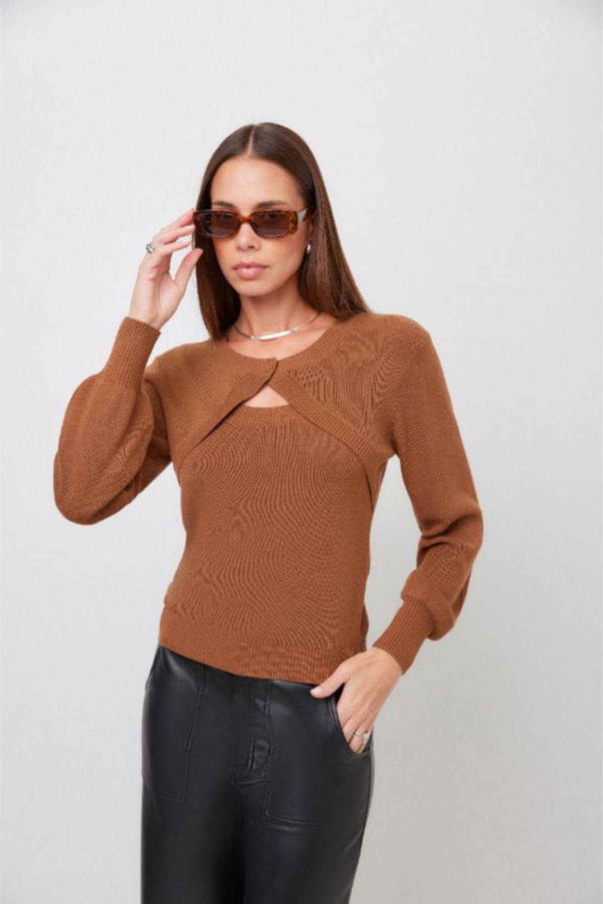 A woman wearing a brown ribbed knit long-sleeve sweater with a keyhole cutout at the chest, paired with black faux-leather pants, against a light gray studio background.