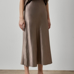 A model wearing a high-waisted, midi-length satin midi skirt in a taupe-brown color, paired with a matching taupe top and brown strappy sandals, set against a neutral studio background.