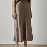 A model wearing a high-waisted, midi-length satin midi skirt in a taupe-brown color, paired with a matching taupe top and brown strappy sandals, set against a neutral studio background.
