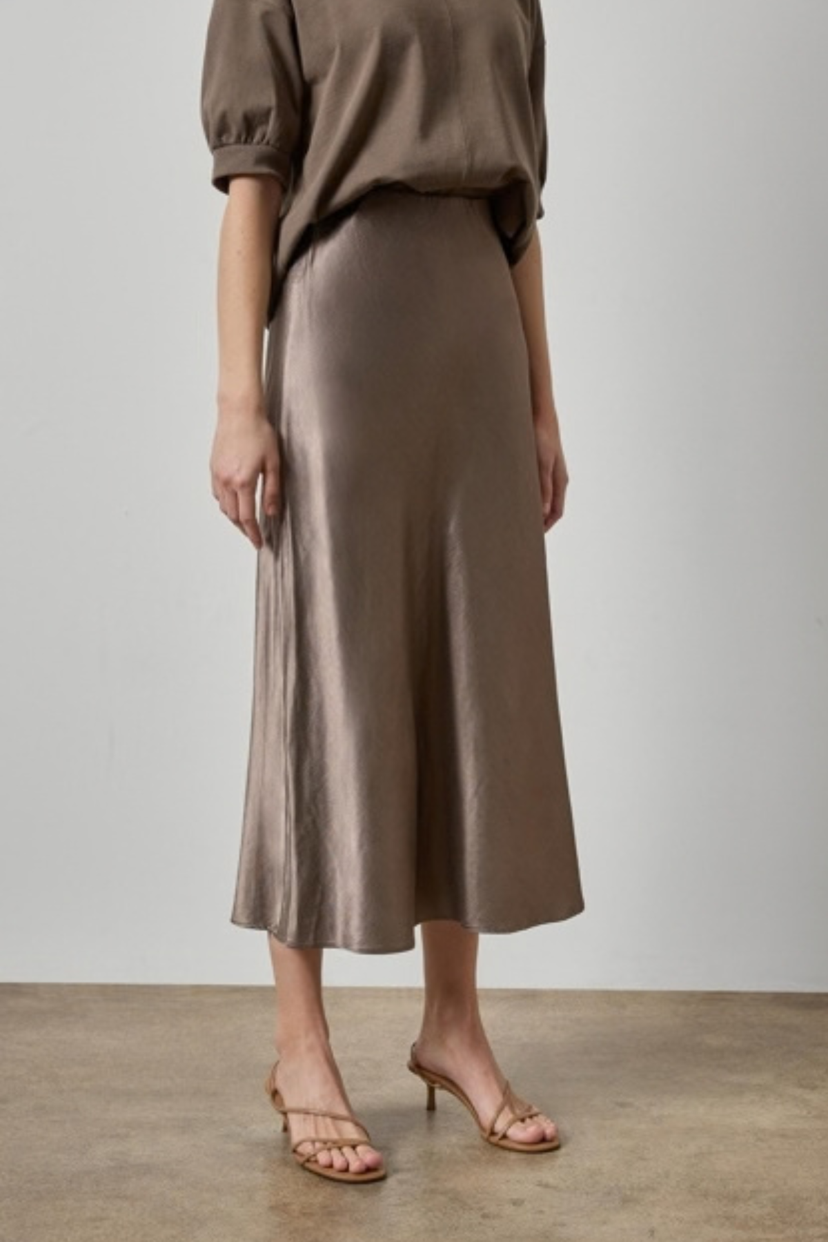 A model wearing a high-waisted, midi-length satin midi skirt in a taupe-brown color, paired with a matching taupe top and brown strappy sandals, set against a neutral studio background.