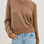 A woman wearing a cropped, long-sleeve V-neck knit sweater in a warm taupe-brown color, paired with light-wash blue jeans, against a plain light gray background.
