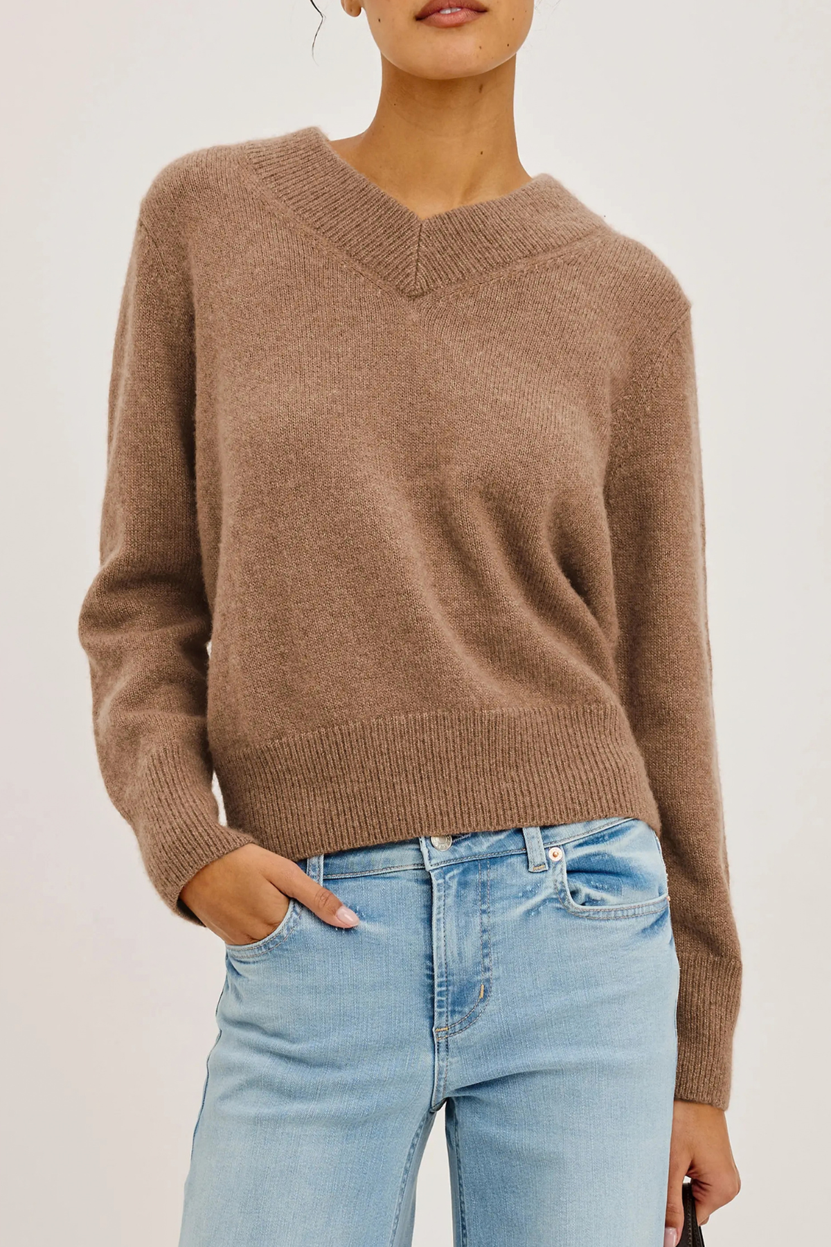 A woman wearing a cropped, long-sleeve V-neck knit sweater in a warm taupe-brown color, paired with light-wash blue jeans, against a plain light gray background.