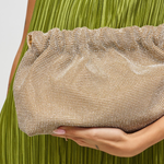 A close-up of a person holding a shimmering champagne clutch bag with a rhinestone-like textured pattern. The person is wearing a pleated olive green dress, and the clutch is positioned against the green fabric.