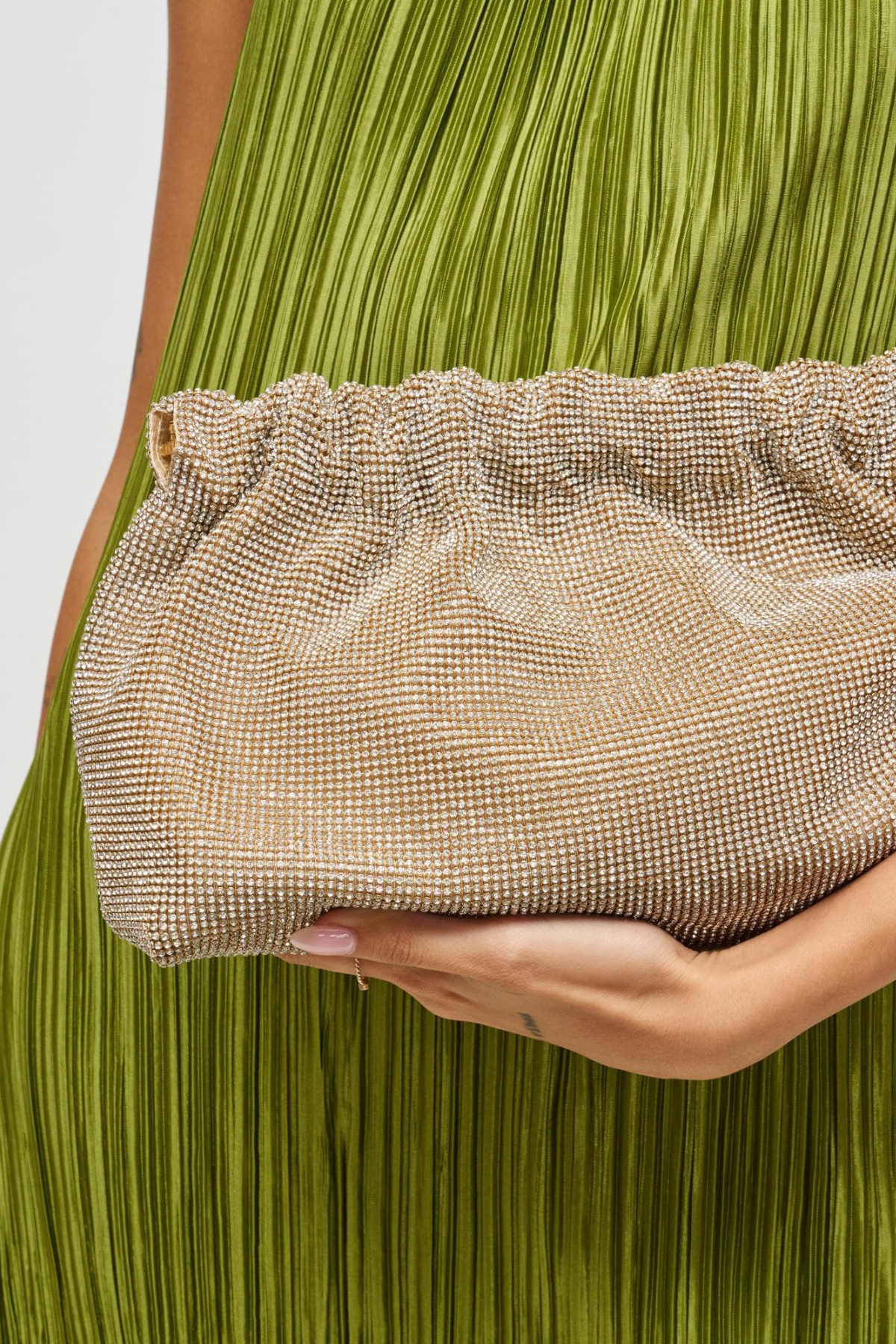 A close-up of a person holding a shimmering champagne clutch bag with a rhinestone-like textured pattern. The person is wearing a pleated olive green dress, and the clutch is positioned against the green fabric.