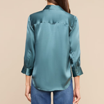 A woman viewed from the back wearing a teal satin button-up shirt and blue jeans against a solid beige background.