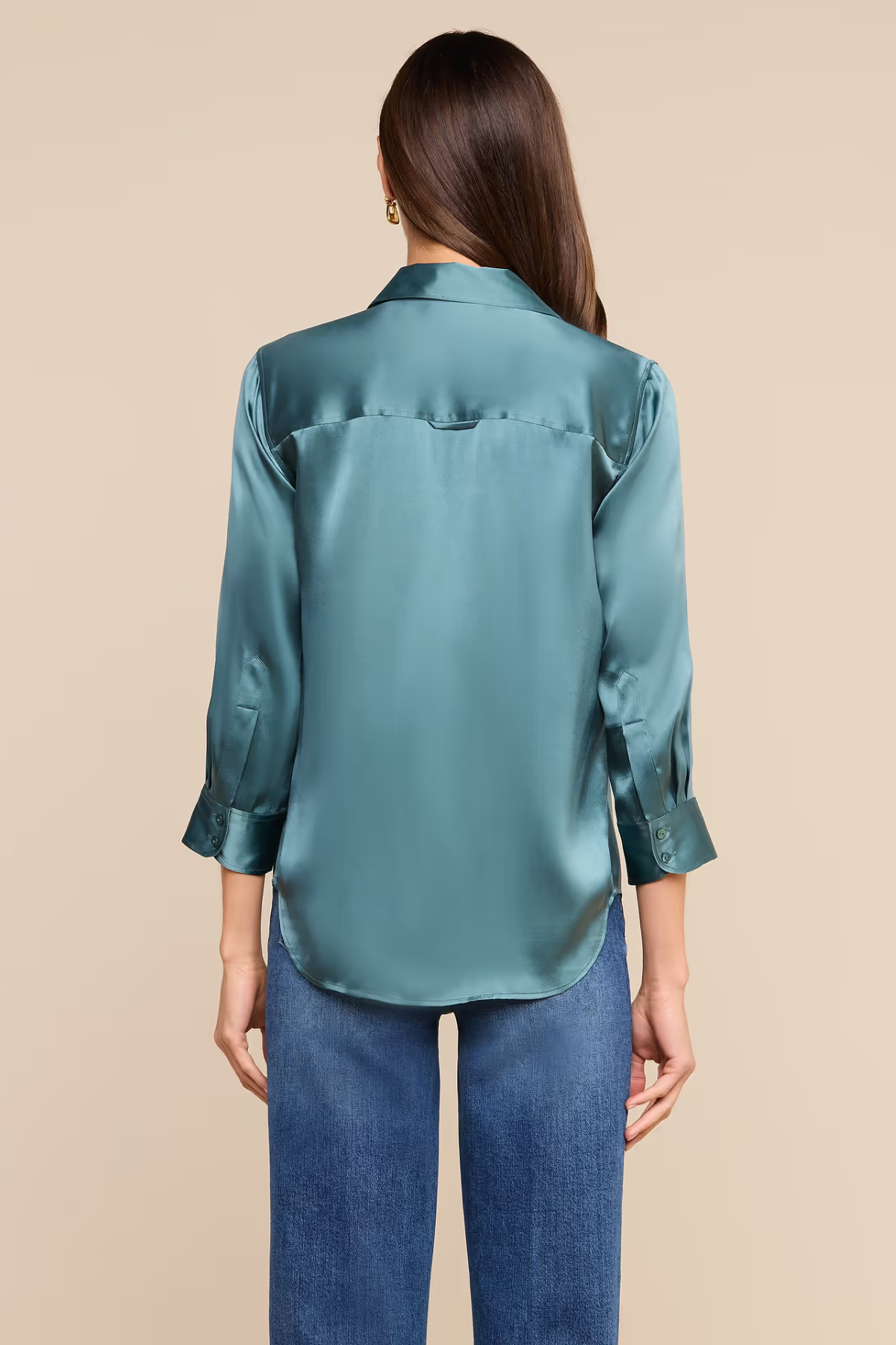 A woman viewed from the back wearing a teal satin button-up shirt and blue jeans against a solid beige background.