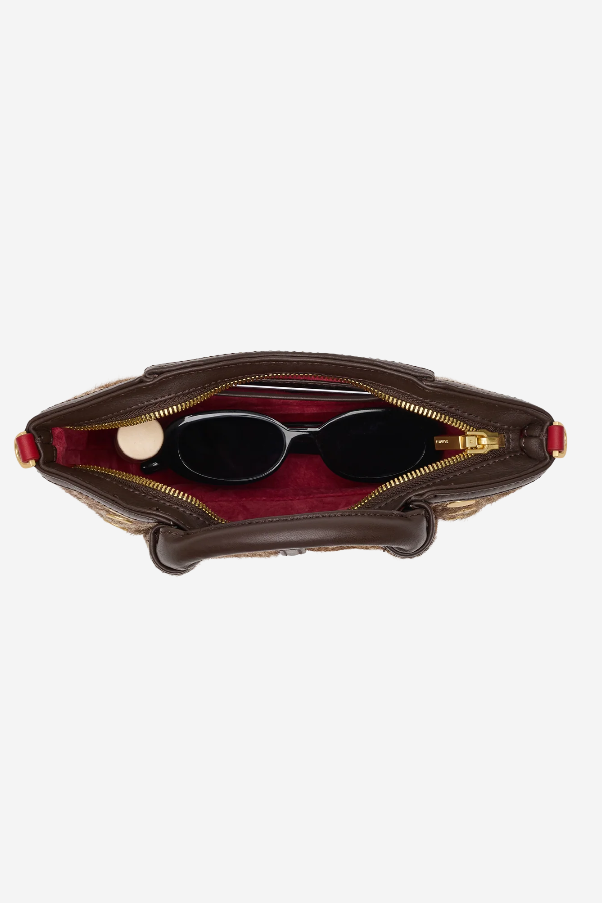 An overhead view of a brown leather handbag with its zipper open, showing a red interior lining and black sunglasses resting inside, set against a plain light gray background.