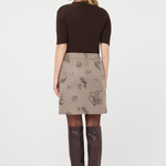 A woman shown from the back wearing a dark brown short-sleeve top, a beige plaid skirt with a paisley pattern, sheer tights, and dark brown knee-high boots, against a plain white studio background.