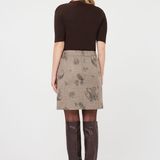 A woman shown from the back wearing a dark brown short-sleeve top, a beige plaid skirt with a paisley pattern, sheer tights, and dark brown knee-high boots, against a plain white studio background.