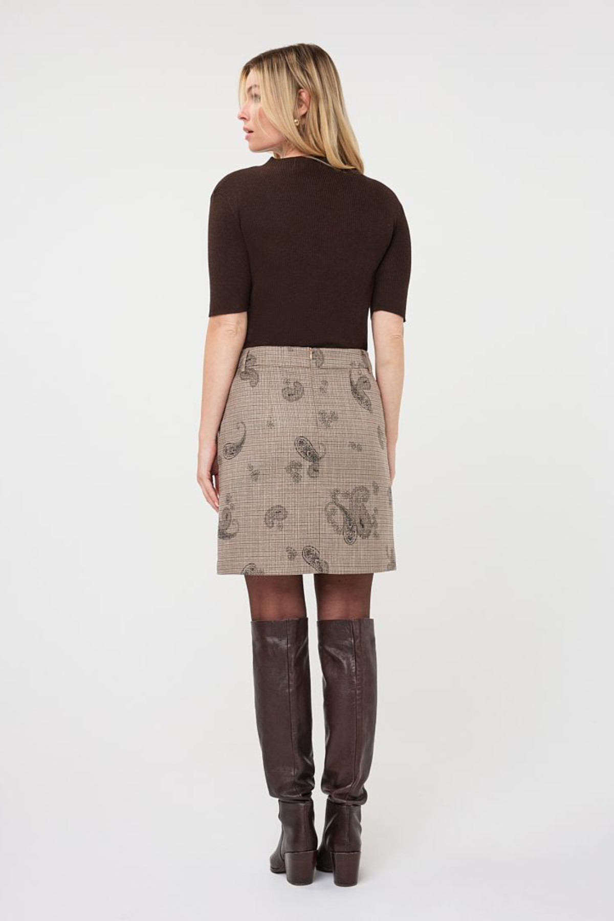 A woman shown from the back wearing a dark brown short-sleeve top, a beige plaid skirt with a paisley pattern, sheer tights, and dark brown knee-high boots, against a plain white studio background.