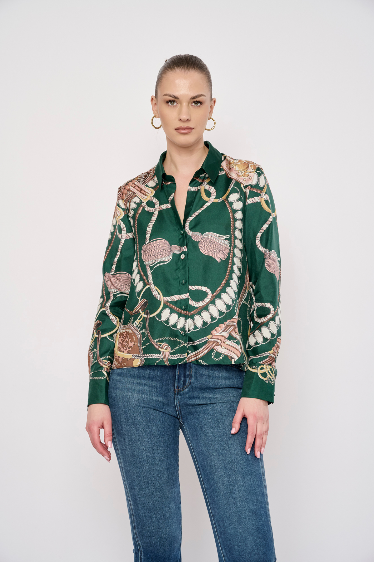 A woman modeling a green long-sleeve button-up shirt with a detailed gold, pink, and white chain-link pattern, paired with blue jeans, against a plain light gray background.