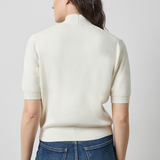 A woman viewed from the back wearing a cream-colored short-sleeve knit mock neck sweater and blue jeans against a light gray studio background.