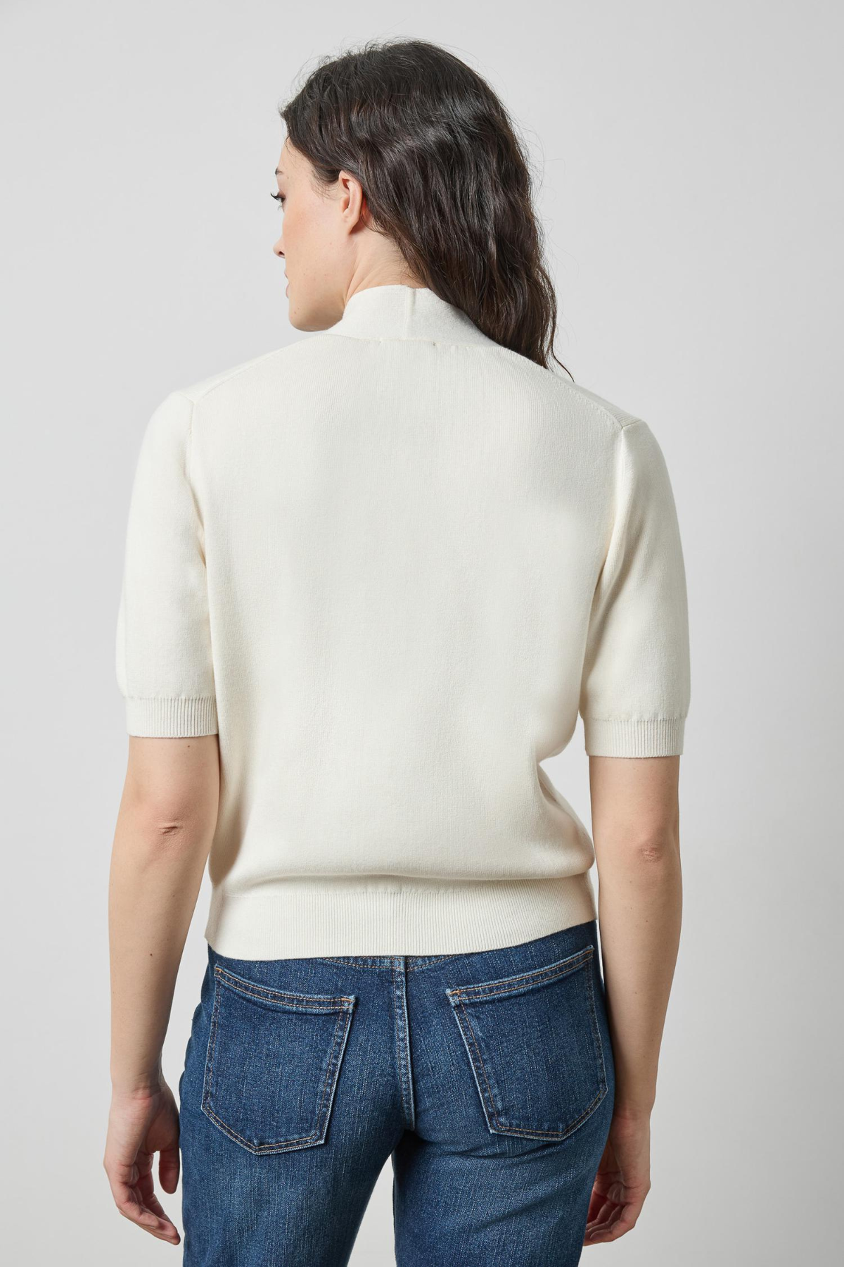 A woman viewed from the back wearing a cream-colored short-sleeve knit mock neck sweater and blue jeans against a light gray studio background.