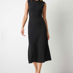 A woman modeling a sleeveless black ribbed midi dress with a high mock neck, standing against a plain light gray wall and concrete floor.