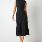 A woman modeling a sleeveless black ribbed midi dress with a high mock neck, standing against a plain light gray wall and concrete floor.