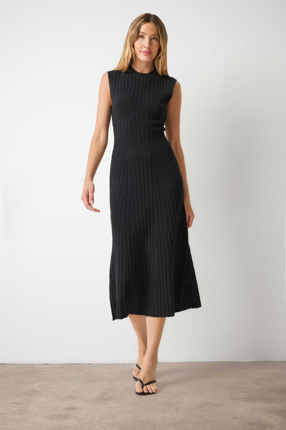 A woman modeling a sleeveless black ribbed midi dress with a high mock neck, standing against a plain light gray wall and concrete floor.