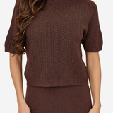 A woman wearing a brown short-sleeve knit sweater with a textured chevron pattern, paired with a matching brown skirt, against a plain light background.