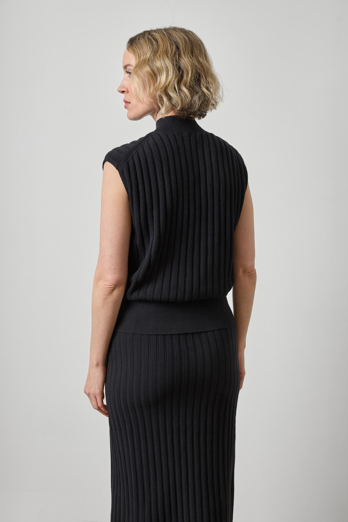 A woman shown from the back wearing a sleeveless black ribbed knit mock-neck top and a matching long black ribbed skirt against a light gray studio background.