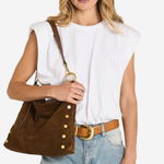 A woman wearing a white sleeveless top, light blue jeans, and a brown belt, carrying a brown suede shoulder bag with gold-tone buttons and hardware.