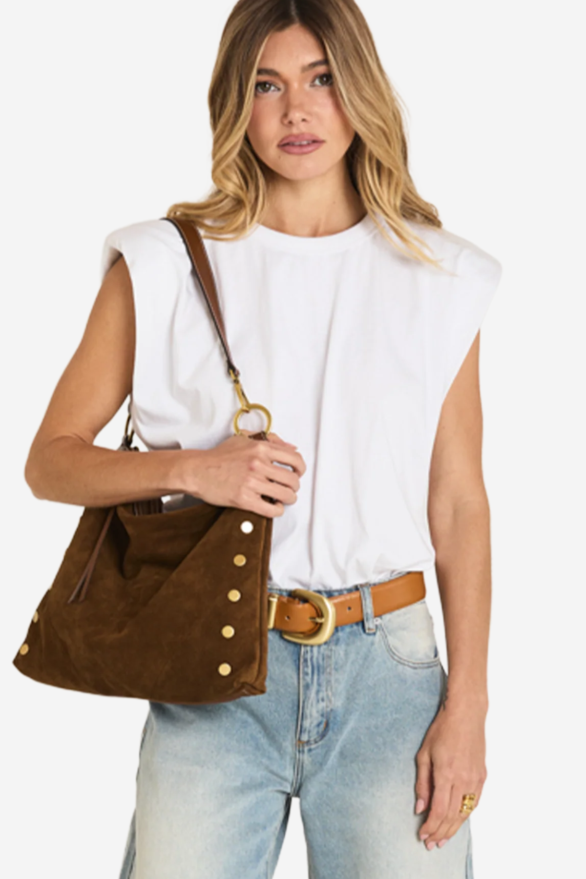 A woman wearing a white sleeveless top, light blue jeans, and a brown belt, carrying a brown suede shoulder bag with gold-tone buttons and hardware.