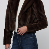 A model wearing a dark brown cropped faux fur jacket over a white turtleneck and blue jeans, shown from the neck down against a light gray studio background.