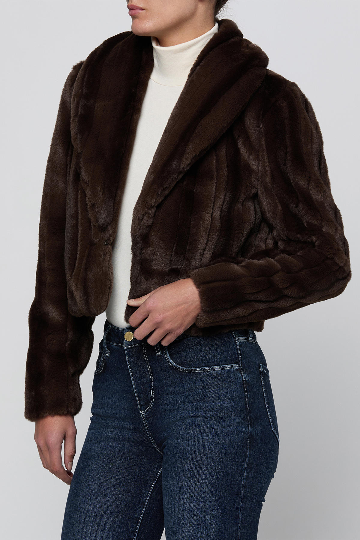 A model wearing a dark brown cropped faux fur jacket over a white turtleneck and blue jeans, shown from the neck down against a light gray studio background.