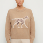 A woman modeling a beige crewneck sweater featuring a large white and gray spotted dog graphic across the chest, paired with light beige trousers, against a plain light gray background.