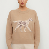 A woman modeling a beige crewneck sweater featuring a large white and gray spotted dog graphic across the chest, paired with light beige trousers, against a plain light gray background.