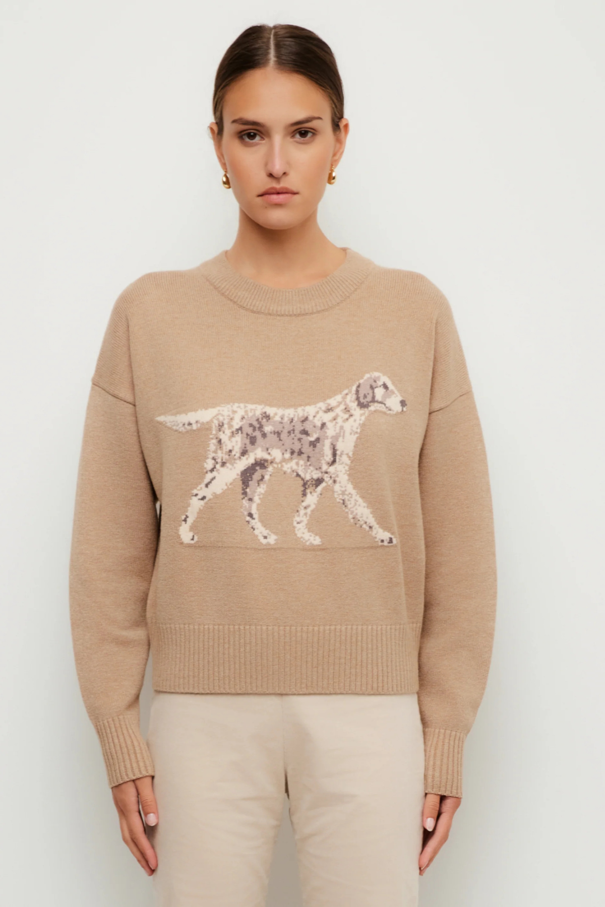 A woman modeling a beige crewneck sweater featuring a large white and gray spotted dog graphic across the chest, paired with light beige trousers, against a plain light gray background.