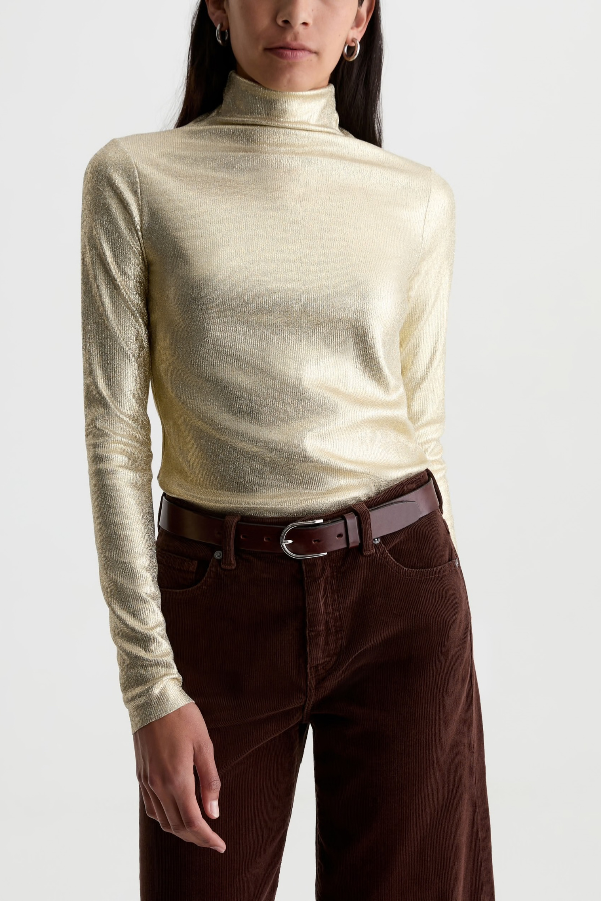 A woman wearing a metallic champagne-gold long-sleeve mock neck top and dark brown corduroy pants with a brown leather belt, photographed against a light gray studio background.