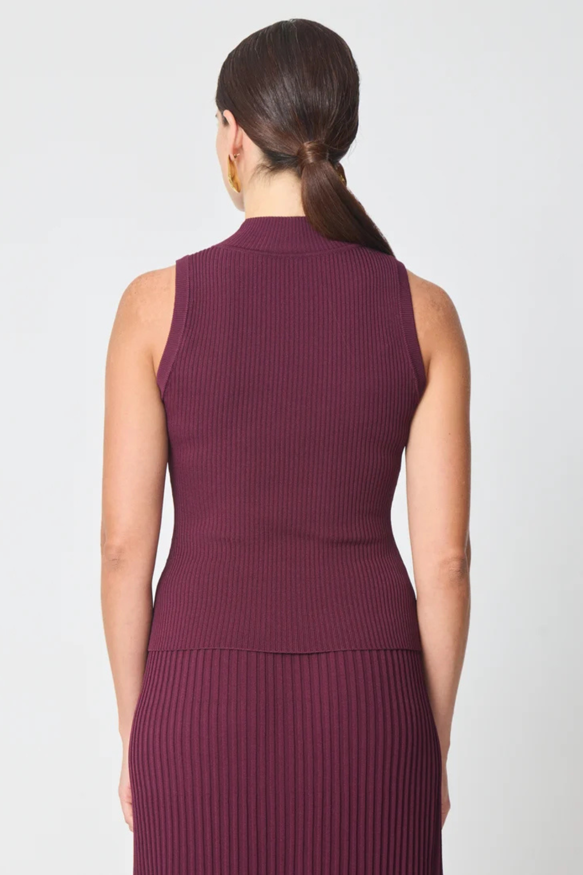 A woman shown from the back wearing a sleeveless ribbed knit top and matching pleated skirt in a deep plum purple color, set against a plain light gray studio background.
