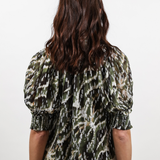 A woman viewed from behind wearing a short-sleeve top with a green, black, and white abstract pattern, paired with blue jeans, against a plain white background.