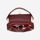 A burgundy leather handbag with gold-tone hardware, shown open to reveal a red interior lining and white sunglasses inside, set against a plain light gray background.