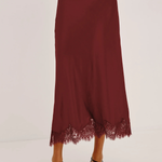 A model wearing a long, dark burgundy midi skirt with a lace hem, paired with black strappy heels and a matching burgundy top, shown against a plain white studio background.