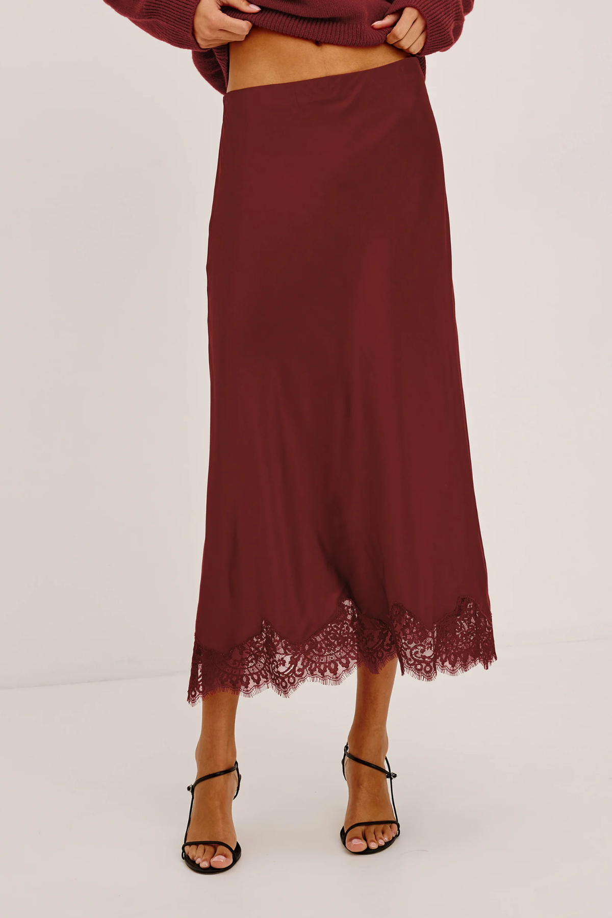 A model wearing a long, dark burgundy midi skirt with a lace hem, paired with black strappy heels and a matching burgundy top, shown against a plain white studio background.