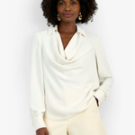 A woman modeling a cream-colored long-sleeve cowl neck blouse paired with textured cream shorts, set against a plain white studio background.