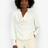 A woman modeling a cream-colored long-sleeve cowl neck blouse paired with textured cream shorts, set against a plain white studio background.