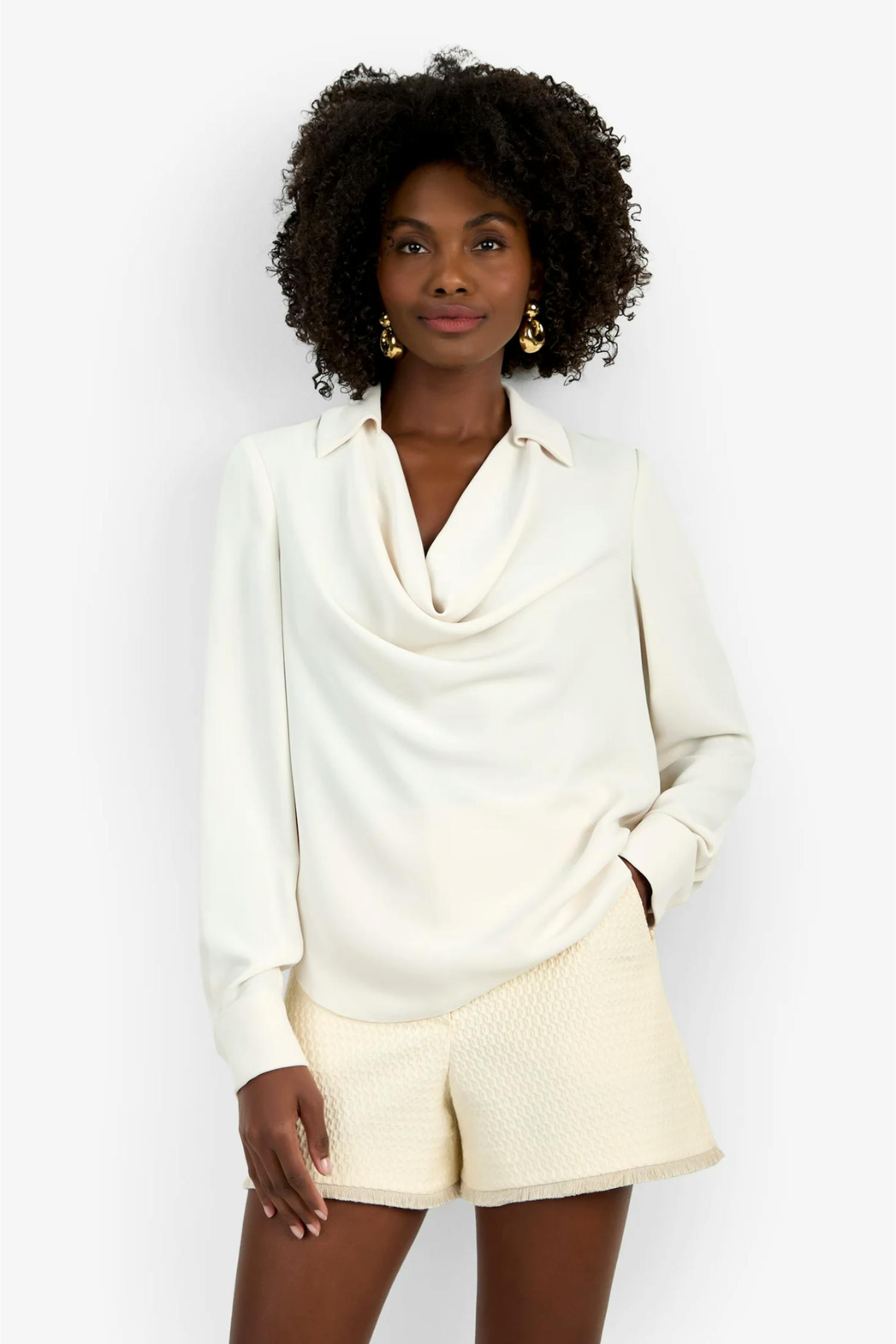 A woman modeling a cream-colored long-sleeve cowl neck blouse paired with textured cream shorts, set against a plain white studio background.